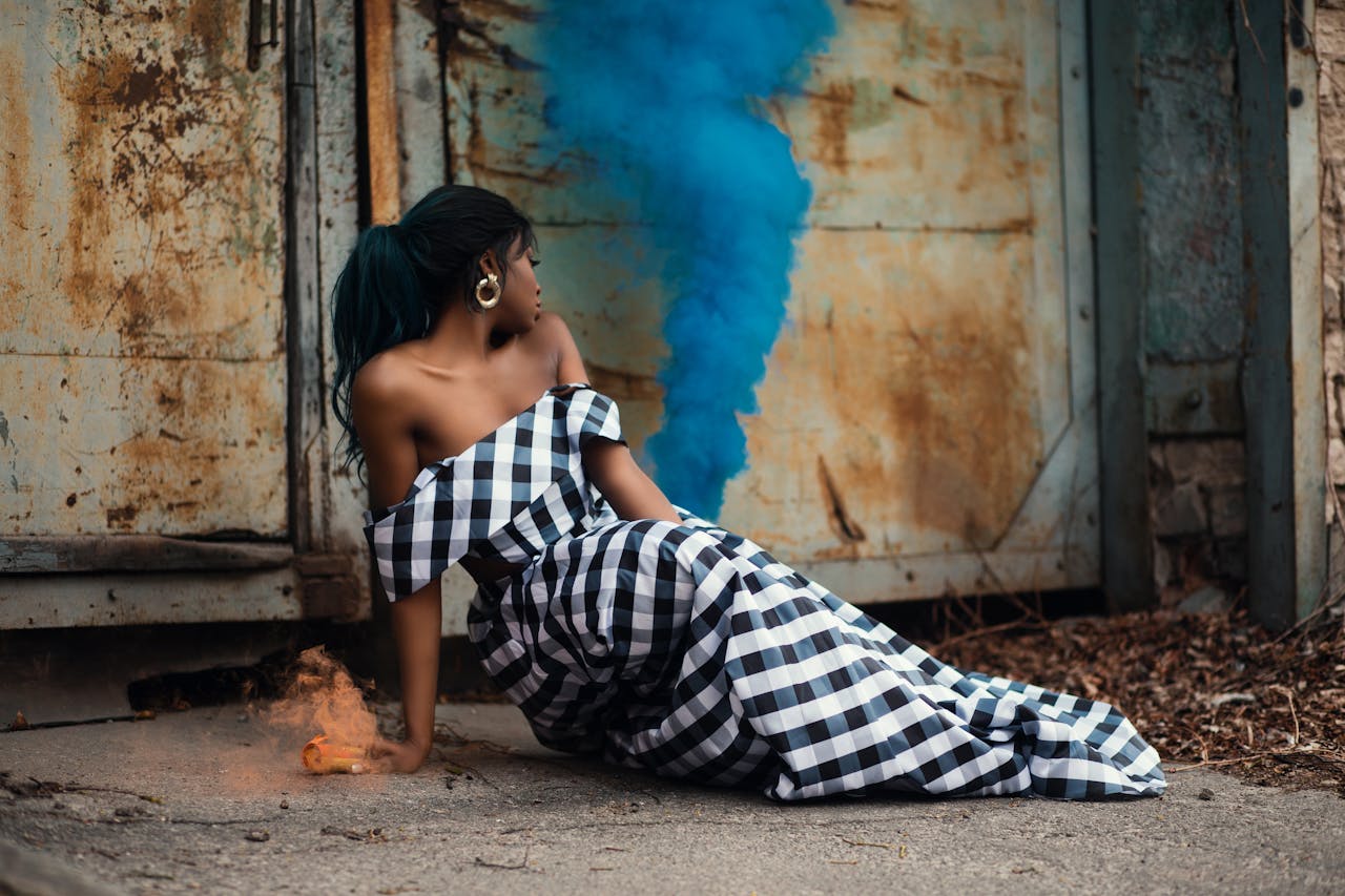 Elegant woman in checkered dress poses with vibrant smoke bomb in rustic urban setting.